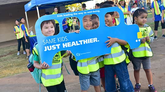 Young students in yellow safety vests holding a cutout board of a school bus